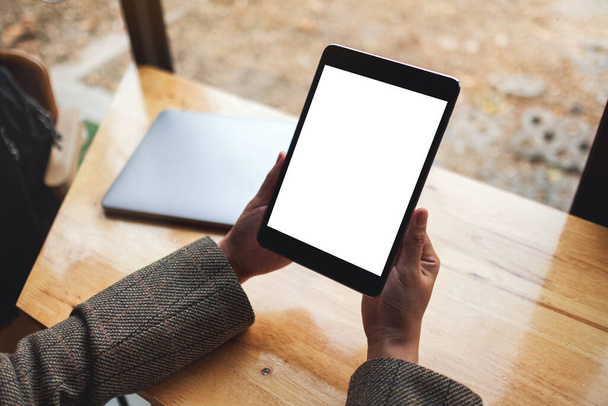 Immagine mockup di una donna che tiene in mano un tablet PC nero con schermo bianco vuoto con laptop su tavolo di legno  - Foto, immagini
