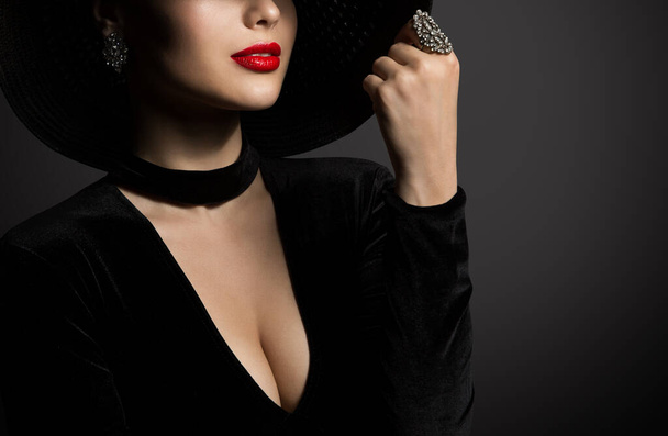 Fashion Woman in Big Hat hidden Face and Red Lips. Sexy Beauty Model in Black Dress Decollete Breast. Elegant Lady Portrait with Necklace over Dark Background showing Ring Jewelry - Fotografie, Obrázek