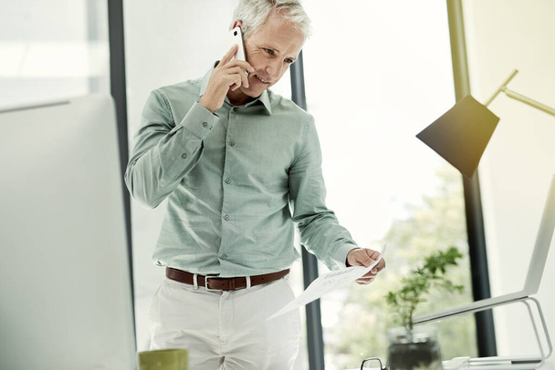 Praten door het contract. Shot van een volwassen zakenman praten aan de telefoon tijdens het werken staan aan zijn bureau in een kantoor. - Foto, afbeelding