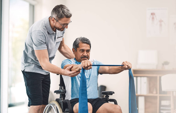 Deze oefening is ook geweldig voor het behoud van een goed evenwicht. Schot van een oudere man in een rolstoel die traint met een verzetsband naast zijn fysiotherapeut.. - Foto, afbeelding