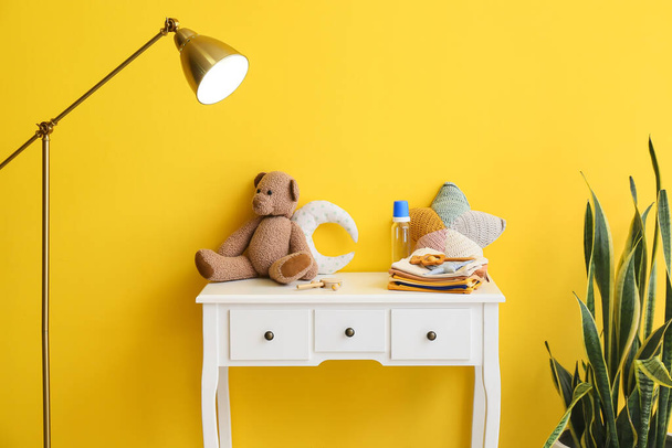 Stapel Babykleidung mit Spielzeug und Flasche auf Tisch neben gelber Wand - Foto, Bild