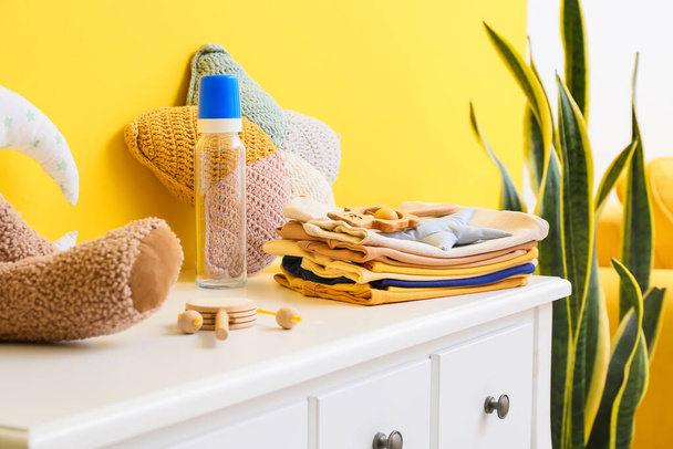 Stapel Babykleidung mit Spielzeug und Flasche auf Tisch neben gelber Wand - Foto, Bild