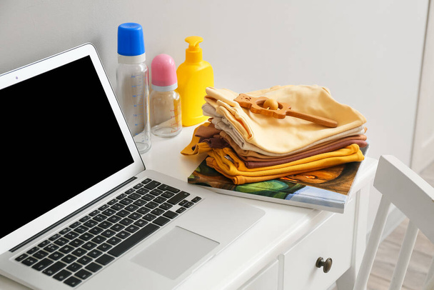 Stack of baby clothes with bottles and laptop on table near light wall - Fotoğraf, Görsel