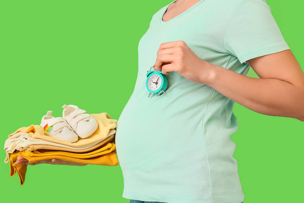 Schwangere mit Stapel Babykleidung und Wecker auf grünem Hintergrund - Foto, Bild