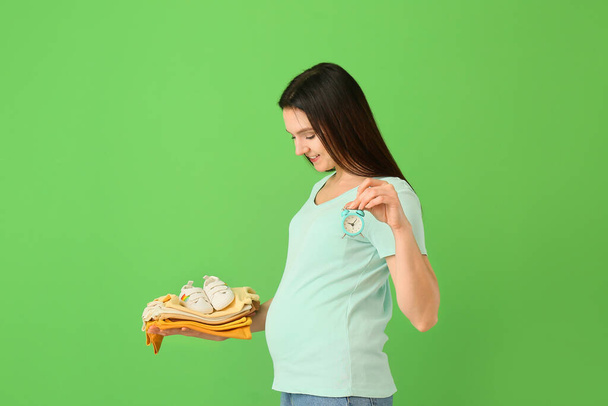 Junge schwangere Frau mit Stapel Babykleidung und Wecker auf grünem Hintergrund - Foto, Bild