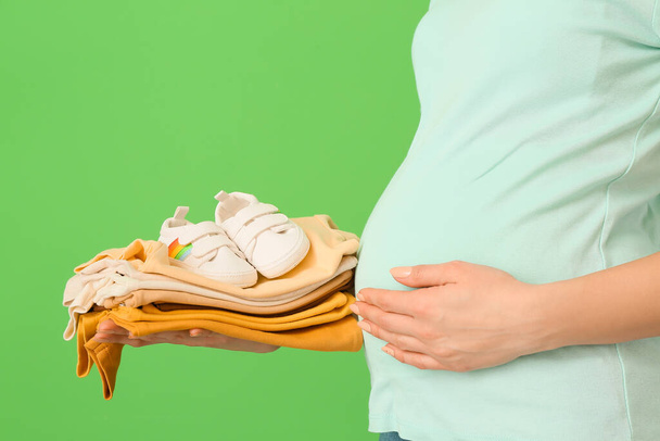 Mulher grávida com pilha de roupas de bebê e botas no fundo verde, close-up - Foto, Imagem