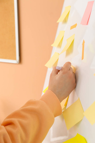 Woman writing on sticky note attached to flipchart near beige wall, closeup - 写真・画像