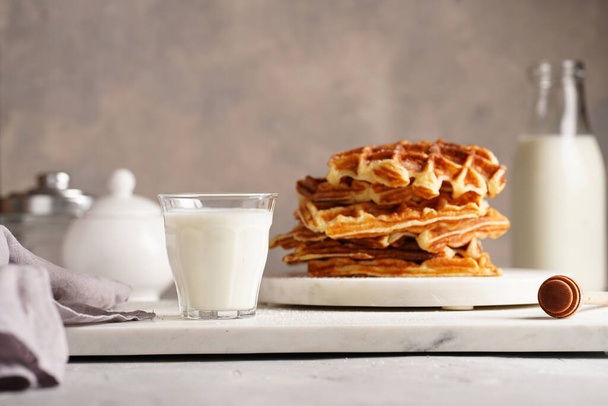 Makea aamiainen - tuoreita kotitekoisia belgialaisia vohveleita hunajalla ja jauhettua sokeria, joka on pinottu valkoiselle pyöreälle marmorilaudalle ja pienelle lasille maidon kanssa - Valokuva, kuva
