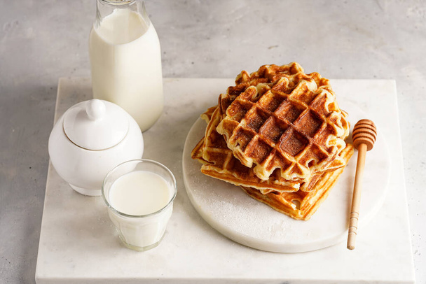 Sladká snídaně - čerstvé domácí belgické vafle s medem a práškovým cukrem naskládané na bílém kulatém mramoru a malé sklenice s mlékem - Fotografie, Obrázek
