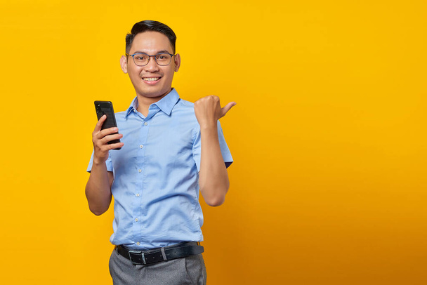 Улыбающийся молодой привлекательный бизнесмен в классической синей рубашке, использующий смартфон и указывающий пальцами в сторону на макет копировального пространства, изолированного на желтом фоне. Достижение бизнес-концепции карьерного богатства - Фото, изображение