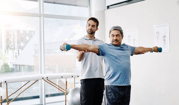 Recorte de un joven fisioterapeuta masculino ayudando a un paciente varón maduro con ejercicios de movimiento en una clínica. - Foto, imagen