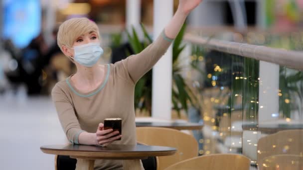 Caucasian adult woman blonde in medical mask mature female visitor sits in cafe at table looks at phone checks time on smartphone looking to side waves invite invitation come here welcome gesture - Footage, Video