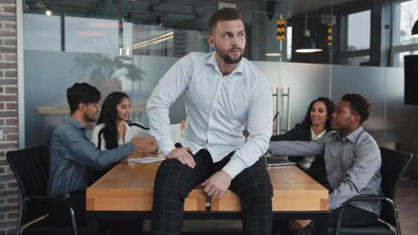 Kaukasische nachdenkliche junge männliche Führer sitzen auf dem Tisch im Konferenzraum selbstbewusst Chef lächelnd Blick in die Kamera zufrieden Teamwork positive freundliche Business-Team diskutieren Projekt Brainstorming - Filmmaterial, Video