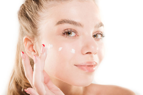 Primer plano retrato de una hermosa joven con piel fresca y crema en su cara mientras posa en el estudio sobre fondo blanco - Foto, Imagen