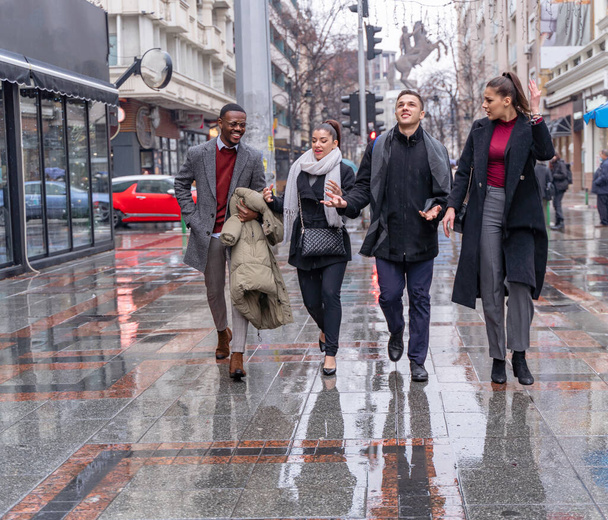 Czterech wielorasowych biznesmenów spaceruje razem po deszczu, rozmawiając o interesach. - Zdjęcie, obraz