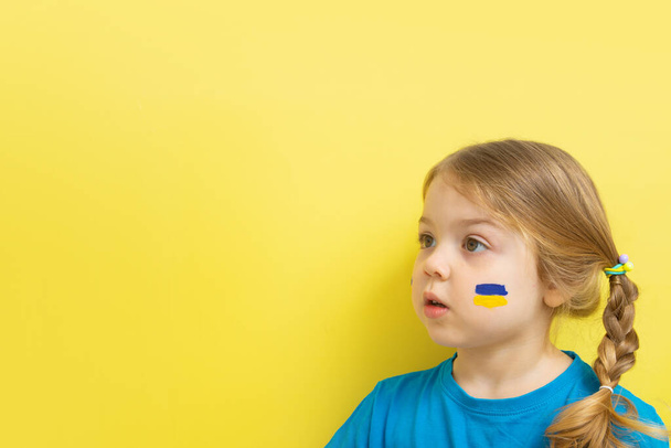 Ragazza di 3 anni con una bandiera gialla e blu dipinta dell'Ucraina sulle guance su uno sfondo giallo con uno spazio di copia. - Foto, immagini