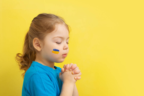 ragazza tenendo le mani giunte in preghiera con i colori giallo e blu della bandiera ucraina sulla guancia - Foto, immagini