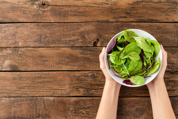Woman hands holding green salad in bowl on rustic wooden background with copyspace. Top view - Photo, image