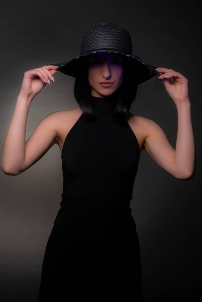 Elegáns lány pózol a kalapjával, miközben fekete ruhát visel, a stúdióban - Fotó, kép