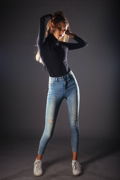 Full-length picture of a beautiful female model posing on dark background in a studio wearing a black blouse, jeans and white sneakers - Fotó, kép
