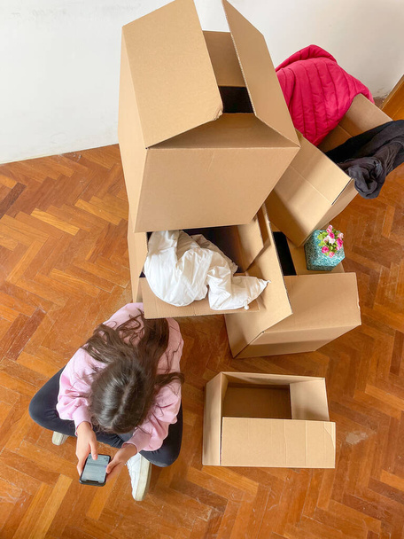Moving to a new home concept. Young girl seated near cardboard boxes along the  parquet floor playing with smartphone, top view - Photo, Image