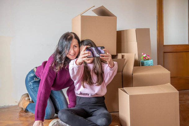Moving to a new home concept. Young girl and her mother seated near cardboard boxes along the  parquet floor taking selfies with smartphone - Foto, Imagem