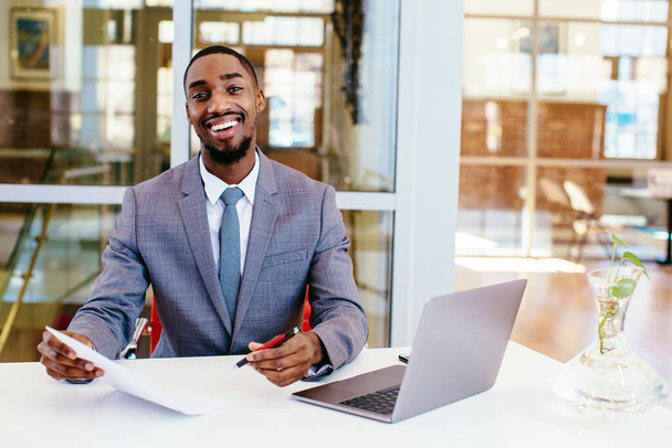 portrét pohledného afrického amerického podnikatele v obleku a kravatě sedícího u stolu a hledícího do kamery - Fotografie, Obrázek