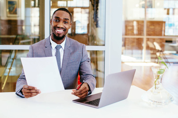 portré egy mosolygó afro-amerikai üzletemberről, aki az asztalnál ül és dokumentumokkal a kezében nézi a kamerát. - Fotó, kép