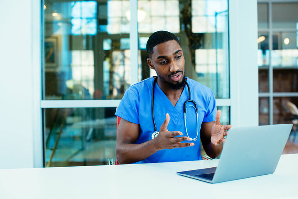 Mavi önlük giyen bir erkek doktorun portresi. İnternette hastayla konuşmak için dizüstü bilgisayar kullanıyor. - Fotoğraf, Görsel
