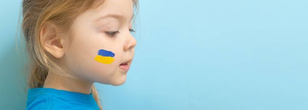 A girl with a painted Ukrainian flag of yellow and blue on her cheeks. Copy space on blue background. - Foto, Imagem