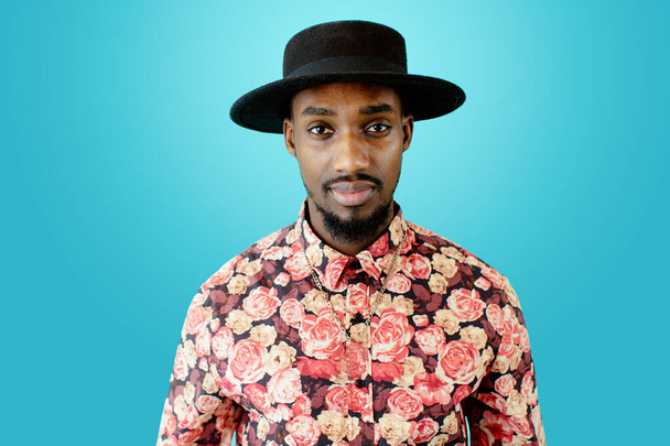 ritratto di un giovane afroamericano in cappello e camicia luminosa posa su sfondo blu - Foto, immagini