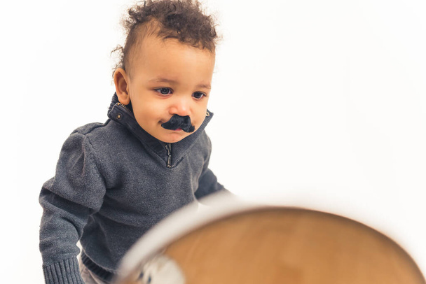 Adorable bebé birracial divertido con bigote falso negro mirándose en el espejo sobre fondo blanco. Captura de estudio. - Foto, imagen