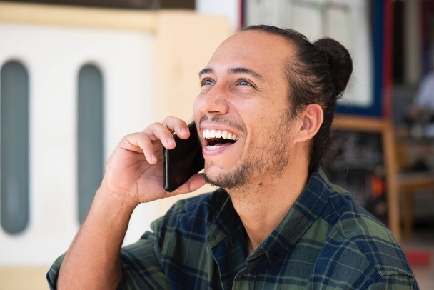 Szczęśliwy człowiek z wyciągniętymi bułeczkami do włosów rozmawiający przez telefon komórkowy na zewnątrz - Zdjęcie, obraz