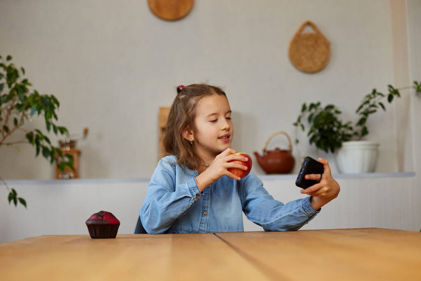 Beautiful young girl filming her blog broadcast about healthy food at the home. Little girl choosing between sweet cake and fresh red apple in , Healthy dieting habit, concept of proper nutrition - Fotografie, Obrázek