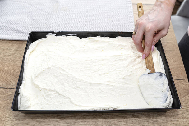 A nő egy kanállal készíti a kevert túrót és a tejszínhabot egy sült kakaós tortán egy sütőlapon.. - Fotó, kép