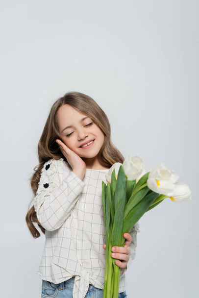 zufriedenes Mädchen mit geschlossenen Augen, das Gesicht berührt, während es Tulpen isoliert auf grau hält - Foto, Bild