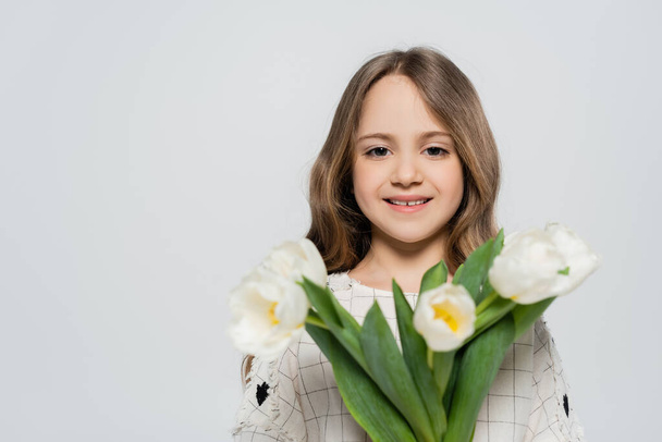 positive girl with blurred bouquet of white tulips looking at camera isolated on grey - 写真・画像