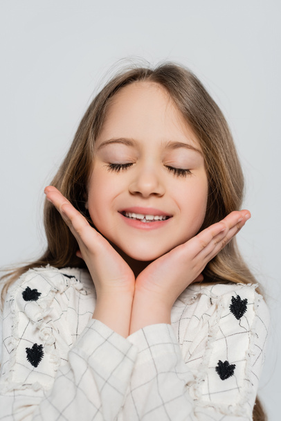 portret van tevreden meisje met gesloten ogen en handen in de buurt gezicht geïsoleerd op grijs - Foto, afbeelding