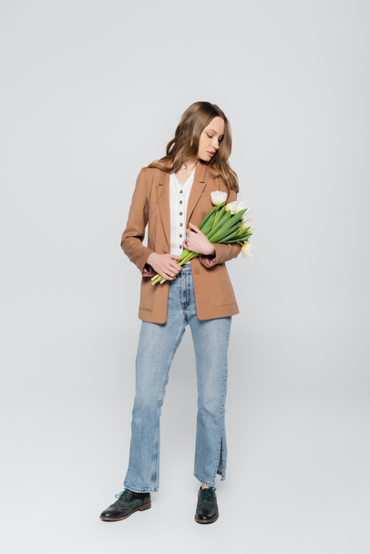 full length view of woman in beige blazer and jeans holding tulips on grey background - Φωτογραφία, εικόνα