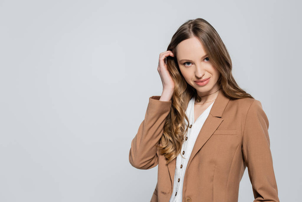 trendy woman in pastel brown blazer looking at camera and touching long hair isolated on grey - Fotó, kép