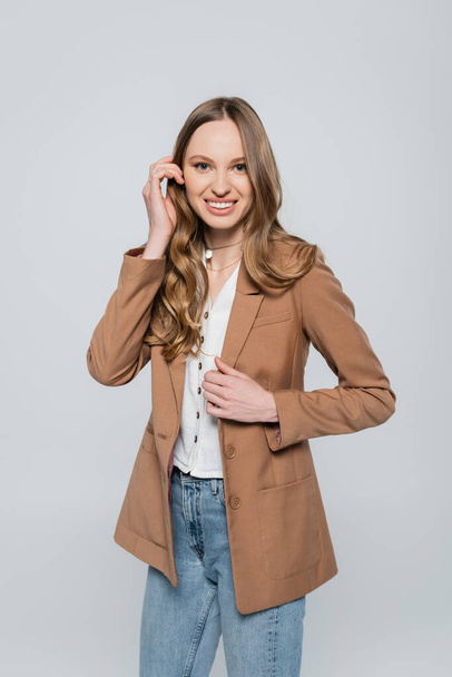joyful woman in beige blazer adjusting hair and looking at camera isolated on grey - Photo, Image