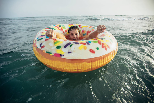 Joyful child on inflatable ring ride on breaking wave. Travel lifestyle, swimming activities. Selective focus - Photo, Image
