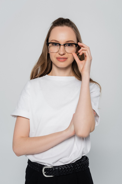 positive woman in white t-shirt adjusting eyeglasses isolated on grey - Foto, Bild