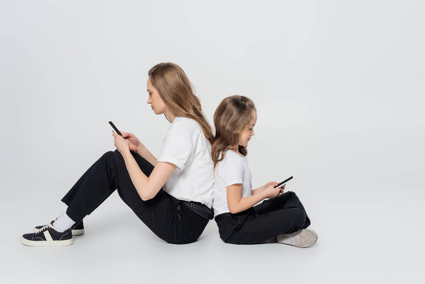 side view of mother and child using smartphones while sitting back to back on grey background - Photo, Image