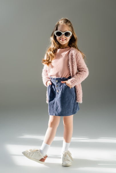 longitud completa de niño feliz en gafas de sol de pie con las manos en los bolsillos en gris  - Foto, Imagen