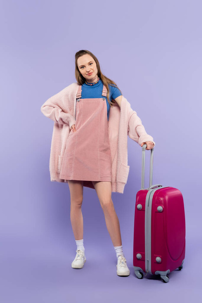 longitud completa de la mujer en rosa vestido general de pie con la mano en la cadera cerca de equipaje en púrpura - Foto, Imagen