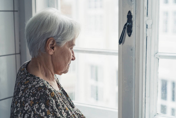 Európai idős nyugdíjas nő az ablak mellett szomorúan néz, és a fiatalságára gondol. - Fotó, kép