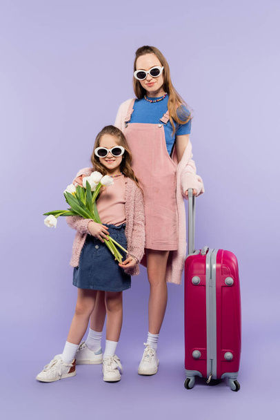 πλήρες μήκος της μητέρας και του παιδιού σε γυαλιά ηλίου κρατώντας λουλούδια και στέκεται κοντά στις αποσκευές σε μωβ - Φωτογραφία, εικόνα