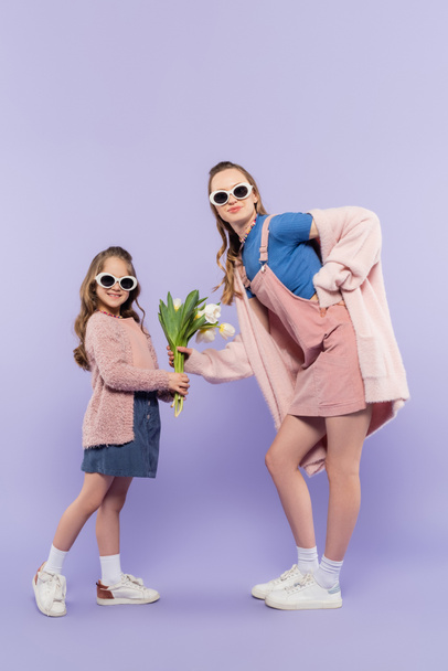 full length of happy girl in sunglasses giving flowers to mother in overall dress on purple - Fotó, kép