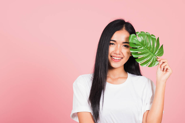 Beauty face. Portrait of Asian beautiful young woman with fresh healthy skin hold green monstera leaf on her face, Tropical Leaves, studio shot isolated on pink background, Skin body care spa concept - Foto, Imagem
